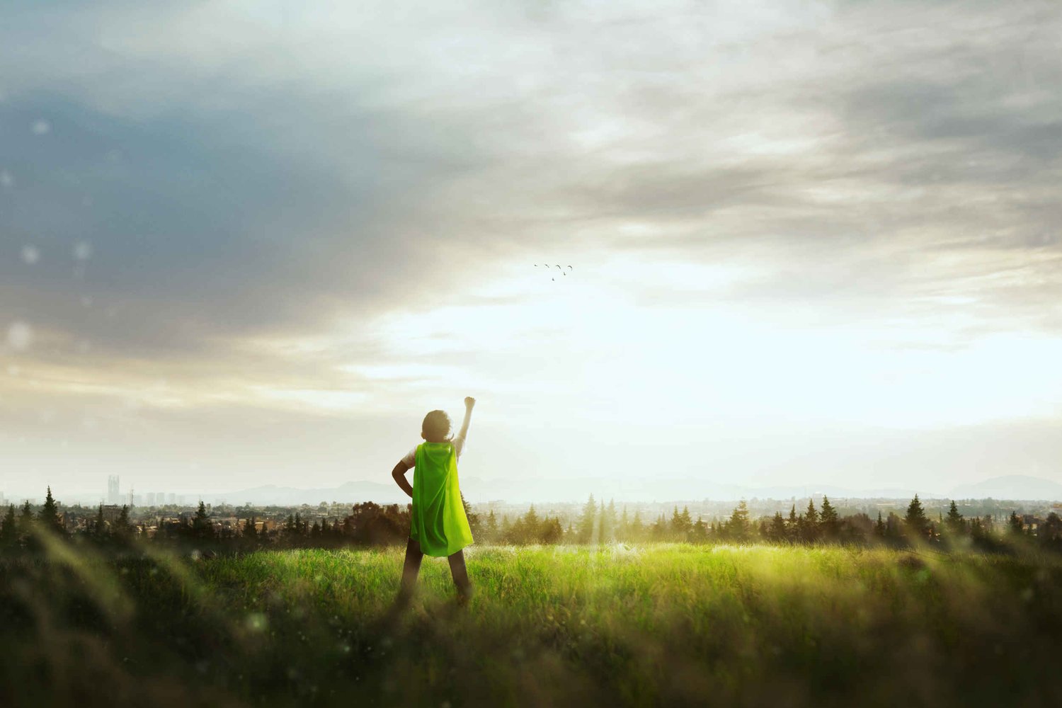 Little boy with a green superhero cape with his back to the camera in the countryside